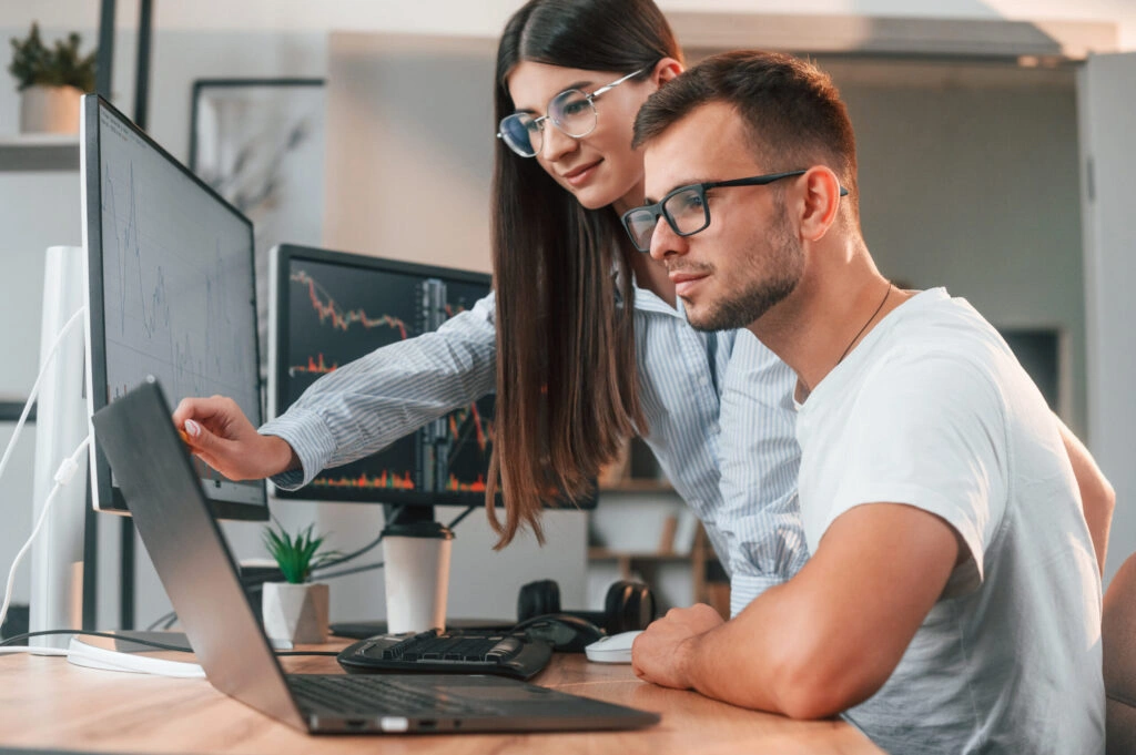 Two people analyzing stock charts on computer screens in a modern office